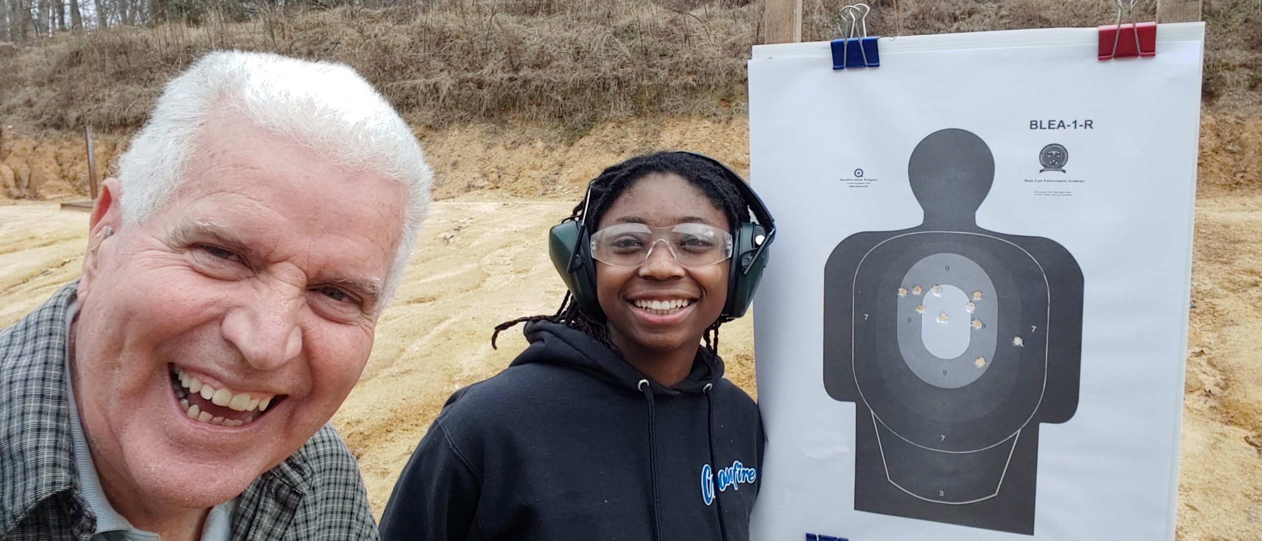 A smiling pistol student wearing eye and hearing protection beside her target